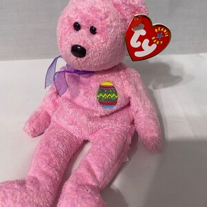 May include: Pink Beanie Baby plush toy with a purple ribbon around its neck. The bear has a black nose and brown eyes. The bear is wearing an embroidered Easter egg on its chest. The tag reads "Beanie Baby".