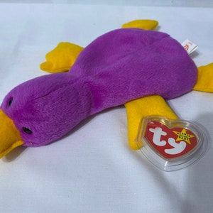 Peut inclure: Jouet en peluche violet et jaune en forme de canard avec une étiquette Ty Beanie Baby attachée. L'étiquette est rouge avec une étoile blanche et le texte "Beanie Baby".