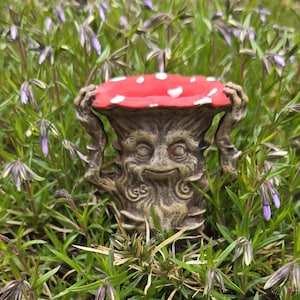 May include: A whimsical garden statue of a mushroom with a smiling face. The mushroom has a red cap with white spots and is supported by a textured, brown base. The statue is surrounded by green grass and purple flowers.