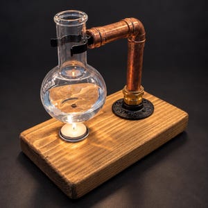 May include: A clear glass round-bottom flask filled with liquid, held by a copper pipe arm attached to a rectangular wooden base. A small lit tea light candle sits on the wood directly beneath the flask, illuminating the liquid from below.