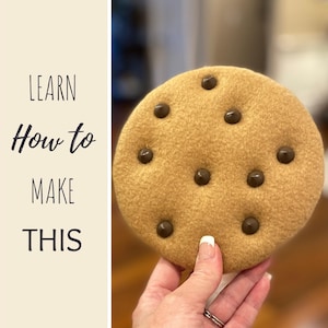 Puede incluir: Una galleta redonda y suave con chispas de chocolate marrón oscuro. La galleta se sostiene en una mano y tiene el texto "LEARN How to MAKE THIS" en el lado izquierdo. La galleta es de color marrón claro.