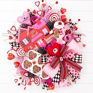 May include: A Valentine's Day wreath with a pink and red color scheme. The wreath features a decorative house with the text "Love Shack", a plush snowman, heart-shaped decorations, and a muffin tin with heart-shaped pastries.