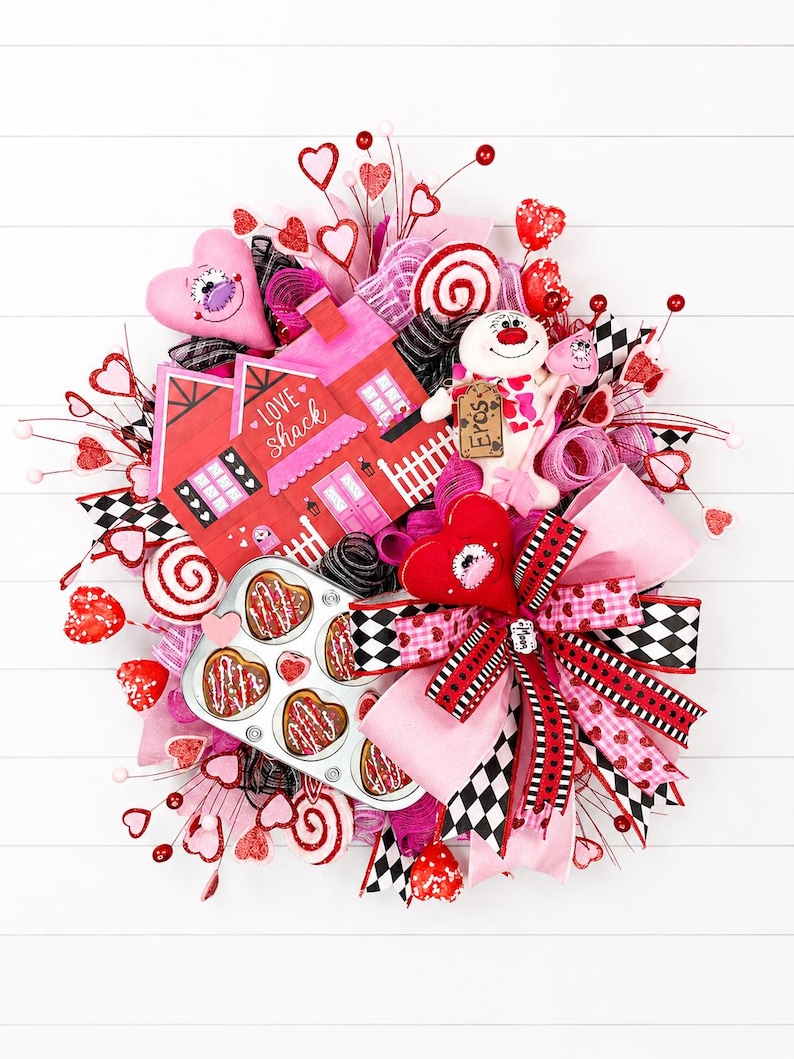 May include: A Valentine's Day wreath with a pink and red color scheme. The wreath features a decorative house with the text "Love Shack", a plush snowman, heart-shaped decorations, and a muffin tin with heart-shaped pastries.