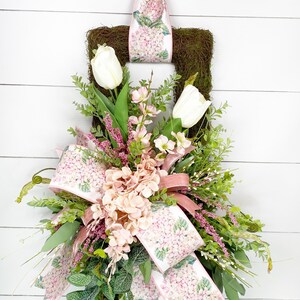 Peut inclure: Une couronne carr&eacute;e en mousse orn&eacute;e de tulipes blanches, d'hortensias roses et de verdure. La couronne pr&eacute;sente un grand n&oelig;ud et un ruban imprim&eacute; de fleurs d'hortensia roses. Un plus petit n&oelig;ud est en haut.