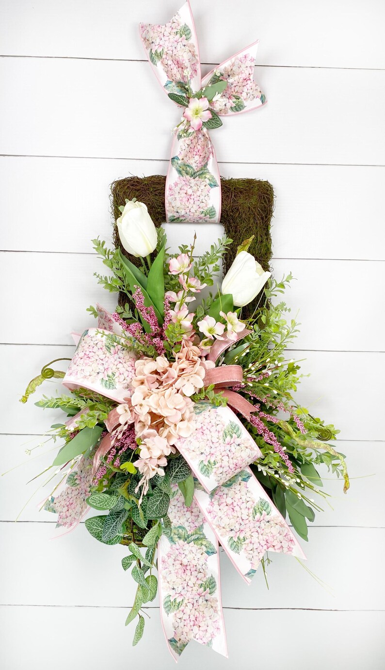 Peut inclure: Une couronne carr&eacute;e en mousse orn&eacute;e de tulipes blanches, d'hortensias roses et de verdure. La couronne pr&eacute;sente un grand n&oelig;ud et un ruban imprim&eacute; de fleurs d'hortensia roses. Un plus petit n&oelig;ud est en haut.