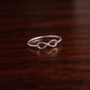 May include: A thin, polished silver-toned ring featuring a central infinity symbol design. The band is smooth and circular, with the infinity loop integrated into the top section of the ring, creating a continuous, symmetrical metal shape.