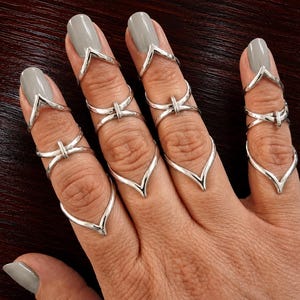 Könnte beinhalten: Ein Set aus silberfarbenen Metallringen an vier Fingern. Jeder Ring hat ein V-förmiges Design am Nagelbett, ein zentrales, überkreuztes Band mit einem vertikalen Steg und ein unteres V-förmiges Band, das zur Fingerbasis hin verläuft.