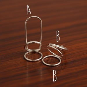 Könnte beinhalten: Silberfarbene Fingerringe. Ein Ring hat ein vertikales Stangendesign, während die anderen ein gestapeltes, offenes Design aufweisen. Die Ringe werden auf einer dunklen Holzoberfläche präsentiert, die ihren metallischen Glanz hervorhebt.