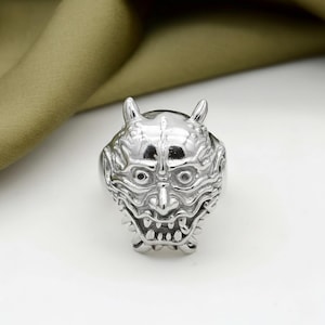 May include: A silver-colored ring featuring a detailed, stylized demon face with horns, eyes, and a wide, toothy grin. The ring has a polished finish and is set against a white background.