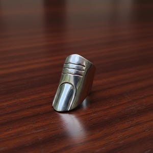 May include: A silver ring shaped like a finger, with a detailed nail and textured lines on the top. The ring is set against a dark wood surface, creating a contrast.