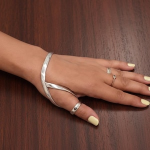 May include: A silver hand bracelet and thumb ring set. The bracelet wraps around the wrist and connects to the thumb ring with a Y-shaped design. Additional rings are worn on the other fingers. The nails are painted yellow.