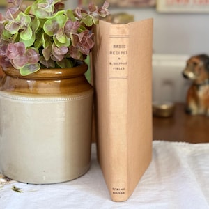 May include: A tan hardback book titled Basic Recipes by M. Sheppard Fidler with Spring Books at the base, positioned next to a two-toned brown and beige ceramic crock filled with a leafy green and purple plant on a white fabric surface.