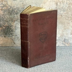 May include: A maroon-covered vintage book, titled "Watch and Clock Making". The book is open, showing aged pages. The title is in gold lettering. The cover features a shield emblem and the name "David Glasgow". The book is from Glasgow.
