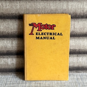 May include: A vintage, yellow book with the title "The Motor Electrical Manual" in red and black. The book is upright against a textured, grey and brown background. The book's cover is slightly worn.