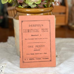 May include: A vintage, rectangular, salmon-coloured softcover book titled Derfel's Geometrical Tests, Standard 5. The cover features black typography, a decorative border, and the text One Penny, along with the publisher information R. J. Derfel, Manchester.