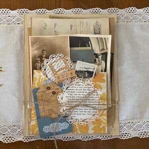 May include: A collection of vintage ephemera including sepia-toned photographs, aged paper documents, and printed text, bundled together with twine. The stack features white paper doilies, a blue cardstock tag, and yellow patterned paper accents.