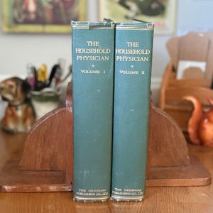 May include: Two vintage teal-covered books, "The Household Physician," Volume I and II, stand in a wooden bookend. The books have gold lettering and are from The Gresham Publishing Co. Ltd. A decorative squirrel figurine is visible.