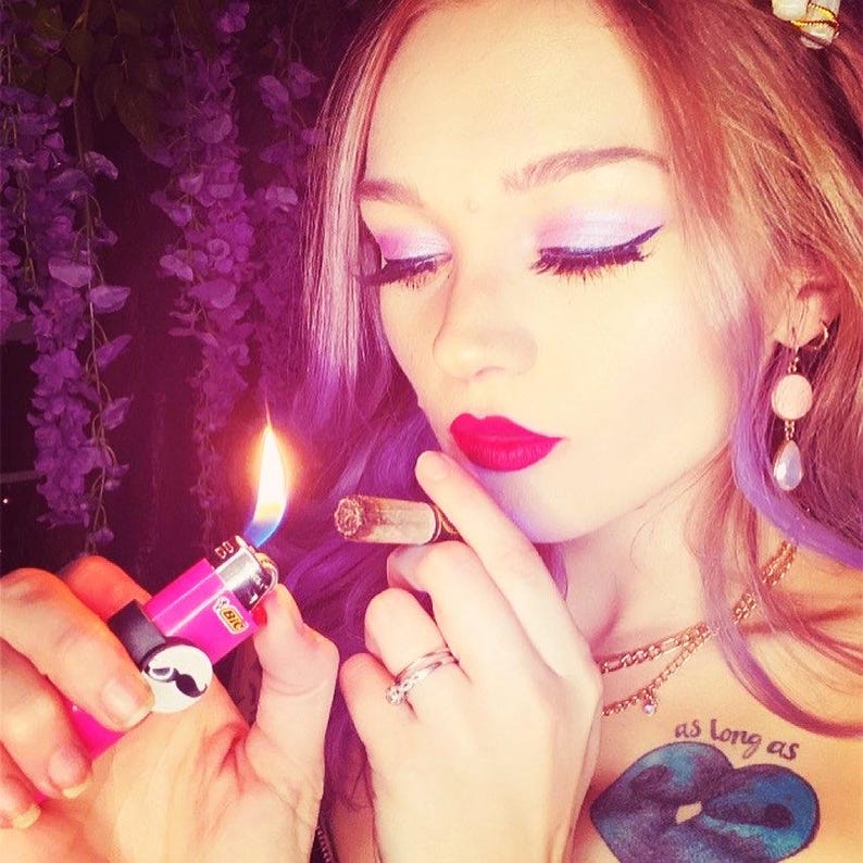 May include: A woman with pink and purple hair is holding a lit cigarette and a pink lighter with a skull and crossbones design. The lighter has the word "Bic" printed on it. The woman is wearing a gold necklace and has a blue tattoo on her chest that says "as long as".