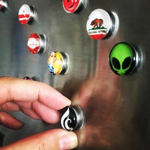 May include: A collection of round refrigerator magnets with various designs. The magnets feature images of a smiley face, a state flag, a bear, an alien, a woman, and a Las Vegas logo. The magnets are attached to a stainless steel refrigerator.