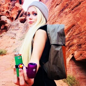 May include: A woman wearing a gray beanie and a brown backpack holds a green lighter and a purple smoking pipe. She is standing in a red rock canyon.