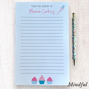 May include: A blue lined notepad with the text "From the bakery of Marna Cakes" at the top. The notepad has a pink and white cupcake illustration at the bottom. The notepad is next to a pen with a colorful confetti design.