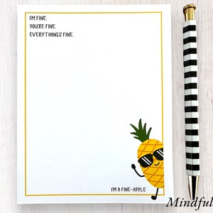 May include: A white notepad with a gold border and a pineapple illustration. The pineapple is wearing sunglasses and has the text "I'M FINE. YOU'RE FINE. EVERYTHING'S FINE." and "I'M A FINE-APPLE" on the notepad. The notepad is on a white wooden surface with a black and white striped pen.