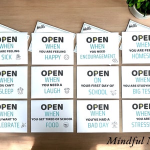 May include: Eight blue and white cards with the text "Open When" followed by a phrase like "You are feeling sick" or "You need encouragement". The cards are on a wooden surface. The text "Mindful Notes" is at the bottom of the image.