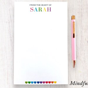 May include: A white notepad with the text "From the heart of SARAH" at the top and a row of colorful hearts at the bottom. The notepad is on a light wood surface with a pink pen next to it. The text "Mindful Notes" is printed below the notepad.