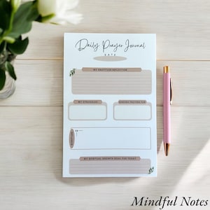 May include: A Daily Prayer Journal notepad with a pink pen. The notepad has sections for gratitude, struggles, prayers, and spiritual goals. The text "Daily Prayer Journal" and "Mindful Notes" are visible.
