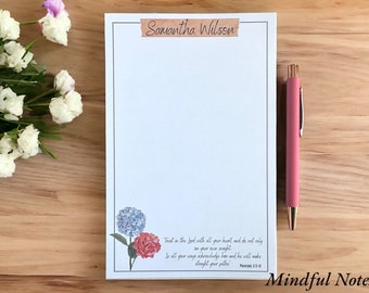 May include: A white notepad with a blue and pink floral design at the bottom. The notepad has a brown paper tape label with the name "Samantha Wilson" written on it. The text "Trust in the Lord with all your heart, and do not rely on your own insight. In all your ways acknowledge him, and he will make straight your paths. Proverbs 3:5-6" is printed at the bottom of the notepad. The notepad is labeled "Mindful Notes".