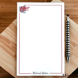 May include: A white notepad with a red border and the word "Ohio" in red lettering. The notepad has a line at the bottom for a name and the words "Mindful Notes" at the bottom right corner. A black and white checkered pen is lying on a wooden surface next to the notepad.