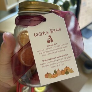 May include: A glass jar with a gold lid, filled with dried orange slices and spices, tied with a burgundy ribbon. A tag reads "Witch's Brew" with instructions for use as a simmer pot or with wine or cider.