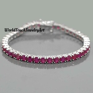 Può includere: Un bracciale d'argento con una fila di pietre preziose rosse scure di taglio ovale. Il bracciale è appoggiato su una superficie grigia. Il testo "WorldTouchJewelryArt" è visibile.