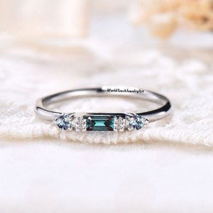 May include: A delicate silver ring featuring a rectangular blue-green gemstone with smaller round gemstones on either side. The ring is displayed on a white lace background.