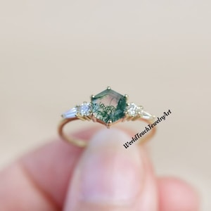 Pode incluir: Um anel de ouro com uma pedra central de ágata musgosa hexagonal, ladeada por duas pedras menores de corte baguette. O anel tem uma banda delicada e está sobre um fundo suave e neutro. O texto "WorldTouchJewelryArt" é visível.