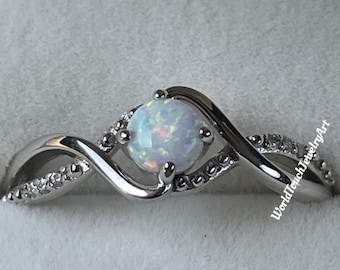 Pode incluir: Um anel de prata com uma pedra de opala branca engastada em uma banda torcida. A banda é adornada com pequenas pedras transparentes. O anel está sobre um fundo branco. WorldTouchJewelryArt está impresso na banda.