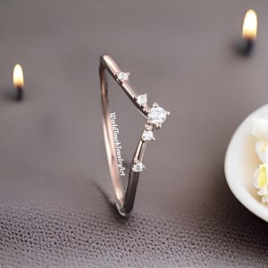 Könnte beinhalten: Ein V-förmiger Ring aus Roségold mit vier klaren, rund geschliffenen Steinen, die entlang einer Seite der Ringschiene gefasst sind. Der Text WorldTouchJewelryArt ist vertikal auf der Innenseite der polierten, schmalen Metallschiene aufgedruckt.