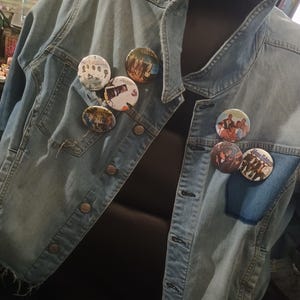 May include: A light blue denim jacket with several round buttons and various band pins attached to the front. The pins feature images of musicians and bands.