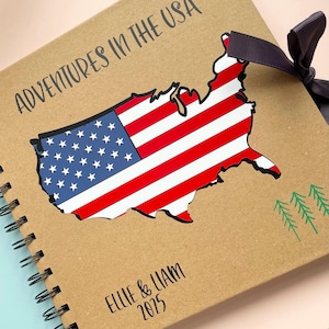 May include: A tan spiral-bound scrapbook with the text ADVENTURES IN THE USA printed at the top. The cover features a map of the United States filled with the American flag pattern, a dark ribbon bow, three green pine tree icons, and ELLIE & LIAM 2025.