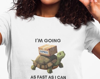 May include: White t-shirt with a graphic of a turtle carrying a package labeled "Express." The text "I'M GOING AS FAST AS I CAN" is printed below the turtle. A person is holding a black smartphone and making a peace sign.
