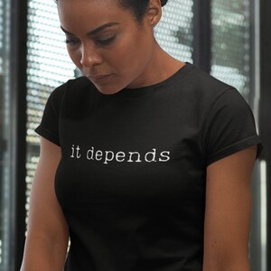 Pode incluir: Uma pessoa vestindo uma camiseta preta com o texto "it depends" em letras brancas, em pé na frente de um laptop e um caderno em uma mesa de madeira.