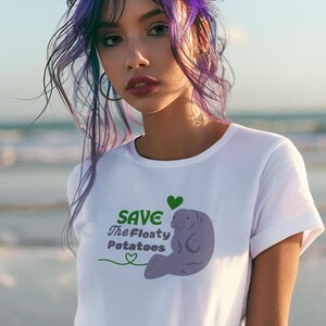 May include: White t-shirt with a graphic of a manatee and the text "SAVE The Floaty Potatoes" in green. A green heart is above the manatee. The shirt is being worn by a person with purple hair.
