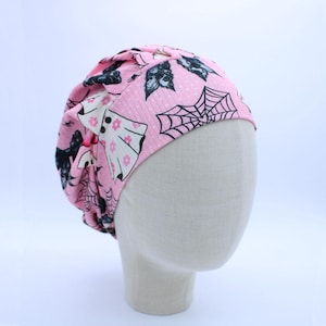 May include: A pink scrub cap featuring a repeating pattern of white ghosts with pink flowers, black spider webs, and black bats. The fabric has a subtle ribbed texture and is gathered at the back to create a rounded, fitted shape on the head.