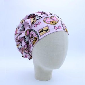 May include: A light pink ribbed scrub cap featuring a repeating pattern of Halloween-themed illustrations including ghosts, jack-o'-lanterns, bats, and pink bows framed in decorative borders. The fabric has a soft texture and a gathered design.