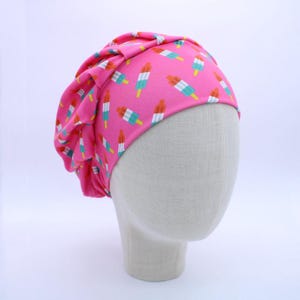 May include: A pink turban-style hat with a repeating pattern of colorful popsicle illustrations. The hat is displayed on a mannequin head against a white background. The popsicles have red, white, blue, and yellow details.