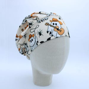 May include: A cream-colored ribbed beanie featuring a repeating pattern of white ghosts wearing orange cowboy hats, playing acoustic guitars, and holding lassos, interspersed with small black spiders across the fabric surface.