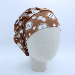 May include: A brown and white patterned hat featuring a repeating snowman design. The hat is made of soft fabric and is displayed on a mannequin head. The snowman pattern includes white snow and black dot eyes and mouth.