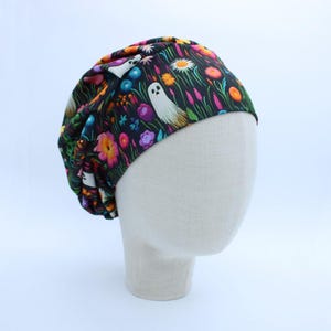 May include: A scrub cap featuring a black background with a colorful pattern of white ghosts, orange, pink, and blue flowers, and green foliage. The fabric is gathered at the top and sides, creating a rounded, pleated shape that fits over the head.
