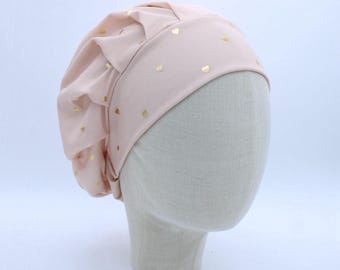 May include: A light pink bonnet featuring a pattern of small gold hearts. The head covering has a wide, smooth band across the front and gathered, pleated fabric at the back, creating a voluminous shape that sits on a neutral-colored mannequin head.