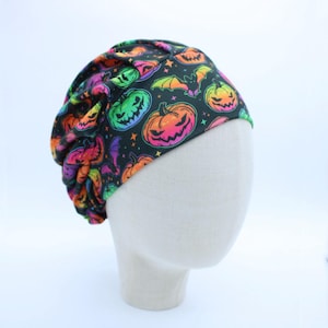 May include: A black scrub cap featuring a vibrant, multicolored pattern of jack-o'-lanterns, bats, and small stars. The fabric displays neon-toned pumpkins in shades of green, orange, and pink, creating a festive Halloween-themed head covering.